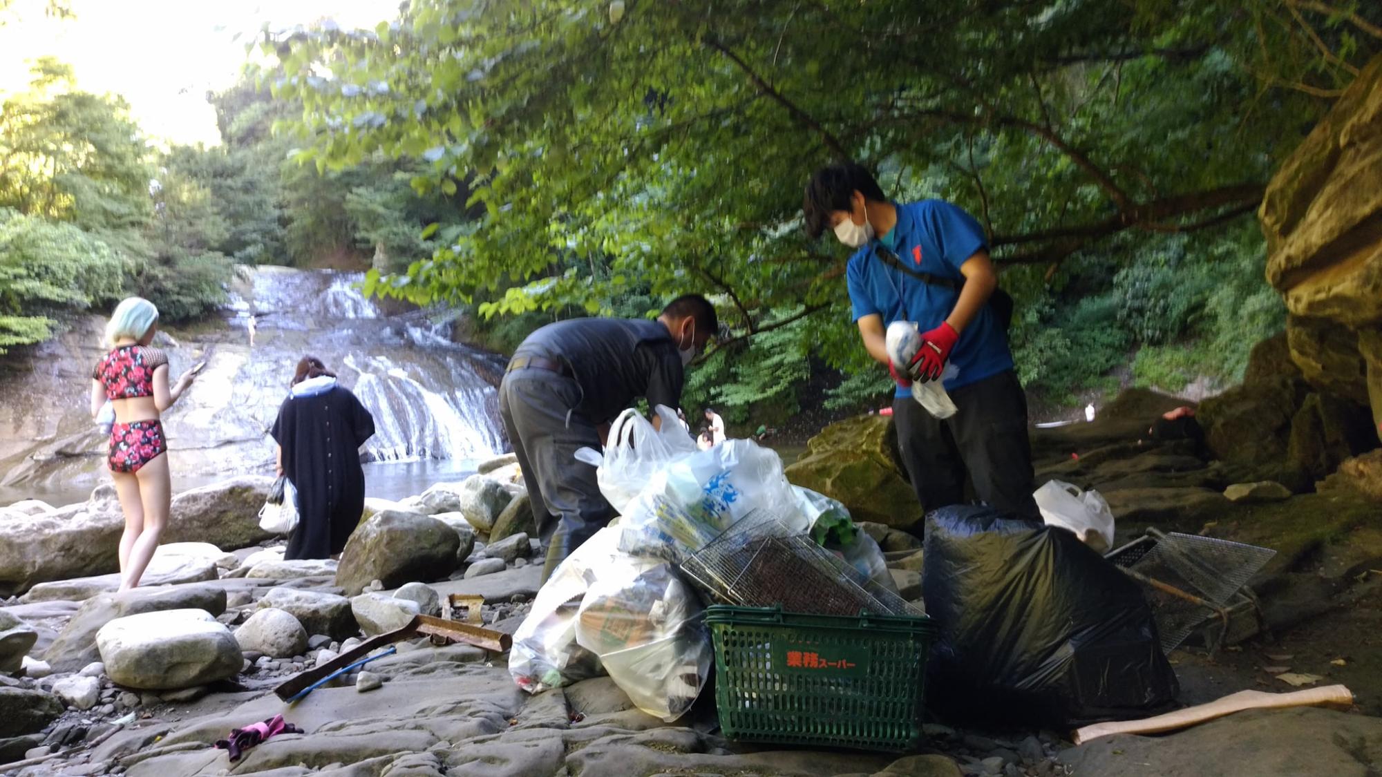 川が流れている横の川辺で4人がゴミを拾い、買い物かごにゴミ袋、その周りにゴミ袋がある写真