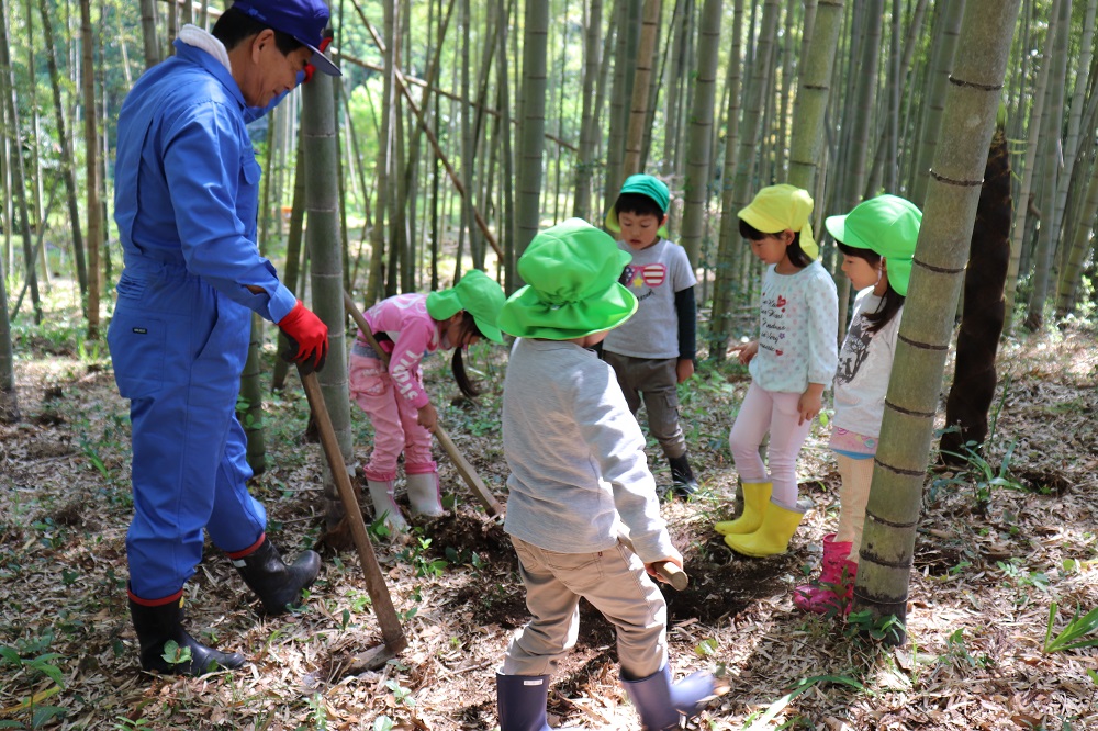 竹林の中にいる、青色の作業着の男性と、帽子をかぶった5人の子どもたちの写真