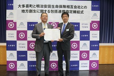 平林町長と明治安田生命保険相互会社千葉本部指導役 菅野様が1つの締結状を2人で持ち、カメラに向かって目線を向けている様子の写真